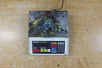 A digital scale with a keypad and display screen is covered in multicolored paint splatters, resting on a wooden surface.