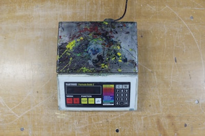 A digital scale with a keypad and display screen is covered in multicolored paint splatters, resting on a wooden surface.