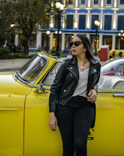 A woman wearing a black leather jacket and sunglasses leans against a yellow vintage car. She stands outside, with an elegant blue and white building in the background. The setting appears to be urban, with other parked cars and trees nearby, creating a stylish and slightly retro atmosphere.