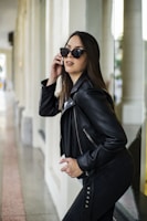 A woman wearing a black leather jacket and sunglasses poses confidently in an outdoor setting beside reflective glass walls and columns. Her long, dark hair flows naturally, and she exudes a modern and stylish vibe.