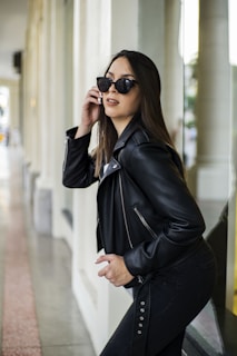 A woman wearing a black leather jacket and sunglasses poses confidently in an outdoor setting beside reflective glass walls and columns. Her long, dark hair flows naturally, and she exudes a modern and stylish vibe.