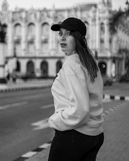 A woman in a casual outfit, including a baseball cap and sweatshirt, stands on a city street with hands in her pockets. The background features an ornate building, suggesting an urban environment with historical architecture.