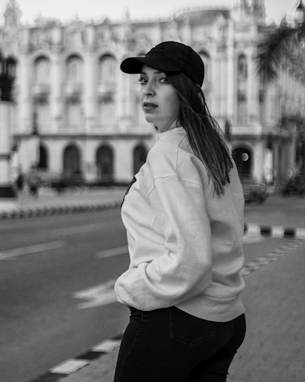 A woman in a casual outfit, including a baseball cap and sweatshirt, stands on a city street with hands in her pockets. The background features an ornate building, suggesting an urban environment with historical architecture.
