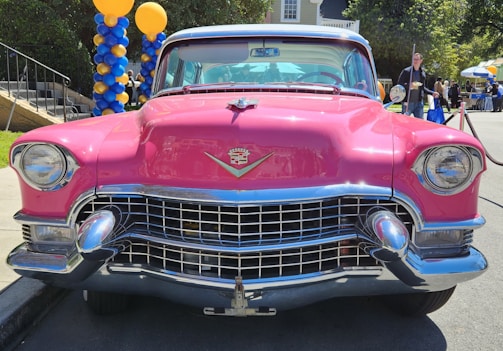 A classic pink vintage car with chrome accents is parked outdoors. The front grille and headlights are prominently displayed. In the background, there are colorful balloon decorations in blue and yellow. A few people are standing nearby, hinting at a public event or gathering.