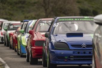 A lineup of race cars is parked in close proximity on a paved road. The focus is on the blue car in the front with the label 'MOTORS BY PINUCCI' on the windshield. The cars behind display a mix of vibrant colors including red and green, indicating a diverse palette typically seen in motor racing.