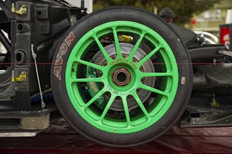 A close-up view of a green racing wheel attached to the frame of a race car. The wheel features a bright green rim with the words 'MG Fury' and 'Racing' inscribed. The tire has 'AVON' branding in white letters. The setting appears to be a garage or workshop, with parts of the car's braking system visible behind the wheel.