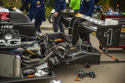 A partially disassembled race car, with various mechanical parts exposed. Three individuals in racing team uniforms are working or standing near the car. There are tools, including wrenches and screwdrivers, scattered on the ground. The background shows a red wheeled toolbox and some green foliage as well as a motorcycle.