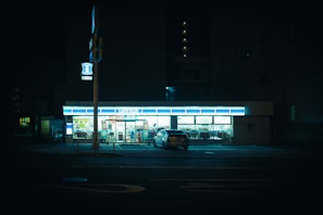A convenience store brightly lit against the darkness of night, with a parked car in front of it. The surrounding area is dimly lit, with minimal activity visible. The store's sign glows prominently in the otherwise subdued streetscape.