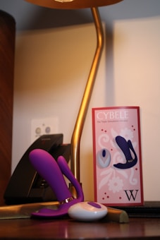 A modern, curved table lamp with a gold finish is positioned next to a purple and white electronic device, possibly a personal massager or novelty item, placed on a wooden surface. A package labeled 'CYBELE' with floral designs and images of the product is nearby. The scene includes a black electronic device, likely a telephone, in the background.