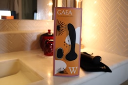 A product box with a sleek design is placed on a bathroom countertop. The box is orange with black and white abstract patterns and features the text 'GAEA The Vibrating Black Dildo.' The countertop is light-colored, possibly marble, with a red decorative jar and a dark fabric case or pouch nearby.