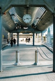 An outdoor walkway with a large clock hanging from a modern, industrial-style overhead structure. Several people are walking on the path beneath the clock, with sunlight casting shadows on the pavement. The background features glass and concrete buildings.