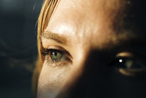 A close-up of a person's eyes, showcasing detailed eyelashes and eyebrows. The lighting casts shadows, creating a dramatic effect on the skin texture.