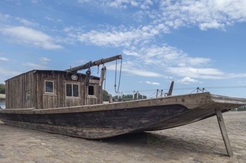 A vintage wooden boat is placed on a cobblestone surface. The boat features a rustic cabin with small windows and a crane-like structure attached to the deck. Overhead, the sky is partly cloudy with patches of blue visible. The background shows hints of a riverbank and some vegetation.