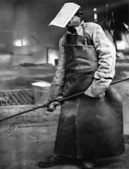A person wearing a protective visor and apron is engaged in an industrial setting. They appear to be handling a long rod, possibly involved in a metalworking or foundry task. The background suggests a workplace with visible equipment and a somewhat dimly lit atmosphere.