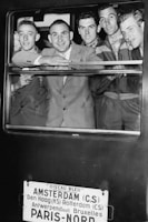 A group of five men dressed in suits and uniforms are smiling and looking out of a train window. Below the window, a sign lists several European cities including Amsterdam and Paris, indicating the train's route.