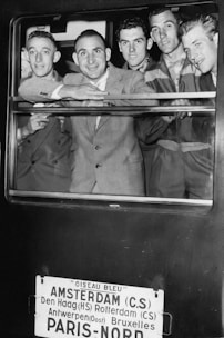 A group of five men dressed in suits and uniforms are smiling and looking out of a train window. Below the window, a sign lists several European cities including Amsterdam and Paris, indicating the train's route.