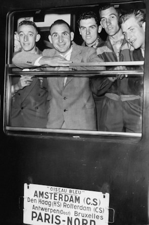 A group of five men dressed in suits and uniforms are smiling and looking out of a train window. Below the window, a sign lists several European cities including Amsterdam and Paris, indicating the train's route.