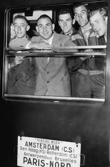 A group of five men dressed in suits and uniforms are smiling and looking out of a train window. Below the window, a sign lists several European cities including Amsterdam and Paris, indicating the train's route.