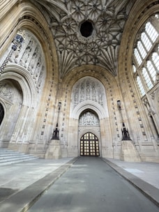 A grand architectural interior features a vaulted ceiling with intricate stonework designs. The walls are adorned with detailed carvings and large arched windows that allow natural light to illuminate the space. A prominent arched doorway with a patterned wooden door stands at the center, framed by statuesque columns and decorative elements.