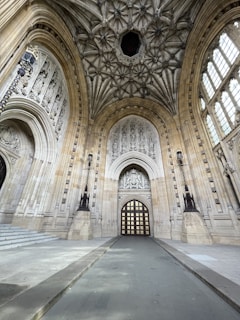 A grand architectural interior features a vaulted ceiling with intricate stonework designs. The walls are adorned with detailed carvings and large arched windows that allow natural light to illuminate the space. A prominent arched doorway with a patterned wooden door stands at the center, framed by statuesque columns and decorative elements.