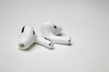 A pair of wireless earbuds resting on a flat, white surface. The earbuds have a sleek design with a glossy finish, featuring small black accents and an ergonomic shape.
