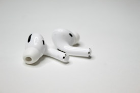 A pair of wireless earbuds resting on a flat, white surface. The earbuds have a sleek design with a glossy finish, featuring small black accents and an ergonomic shape.