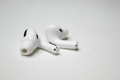 A pair of wireless earbuds resting on a flat, white surface. The earbuds have a sleek design with a glossy finish, featuring small black accents and an ergonomic shape.