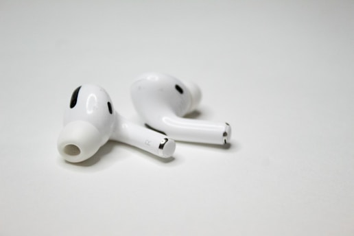 A pair of wireless earbuds resting on a flat, white surface. The earbuds have a sleek design with a glossy finish, featuring small black accents and an ergonomic shape.