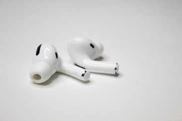 A pair of wireless earbuds resting on a flat, white surface. The earbuds have a sleek design with a glossy finish, featuring small black accents and an ergonomic shape.