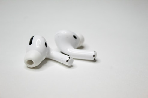 A pair of wireless earbuds resting on a flat, white surface. The earbuds have a sleek design with a glossy finish, featuring small black accents and an ergonomic shape.
