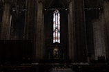 A dimly lit interior of a cathedral with towering stone columns on either side. A large, intricate stained glass window dominates the center, allowing colorful light to filter through the dark space. Below the window, there is an altar with religious artwork and statues.