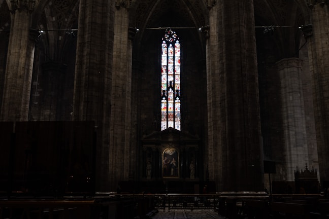 A dimly lit interior of a cathedral with towering stone columns on either side. A large, intricate stained glass window dominates the center, allowing colorful light to filter through the dark space. Below the window, there is an altar with religious artwork and statues.