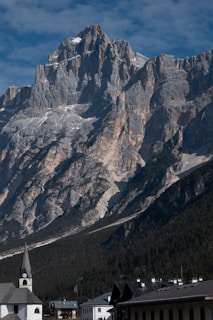 A majestic mountain range with rugged, steep cliffs rising dramatically against a clear, blue sky. The foreground features a small town with buildings and a distinct church steeple. The lower slopes of the mountains are covered in dense, dark green forest.