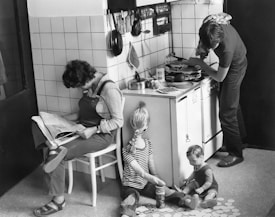 A woman is sitting on a chair reading a magazine in a tiled kitchen. She is leaning against a wall near a stove where a man is cooking. Two children are sitting on the floor, playing with what seems to be large paper circles. Pots, pans, and kitchen utensils are hanging on the wall above the stove.