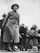 A woman is passionately speaking, wearing a coat and hat, surrounded by a group of attentive people. The background is a plain, cloudy sky, giving a sense of an outdoor gathering or protest.
