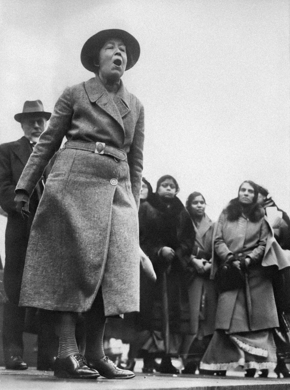 a woman in a long coat and hat standing in front of a group of people