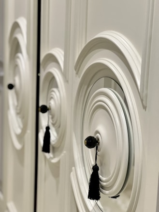 A detailed and close-up view of an ornate white cabinet door featuring circular designs in relief, with black tassel handles hanging from the center knobs.