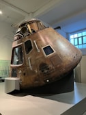 A bronze-colored, weathered spacecraft module displayed in a museum setting. It has multiple windows and visible metal rivets, indicating its durable construction. The surroundings include a smooth, light-colored floor and a large window, contributing to the well-lit environment.