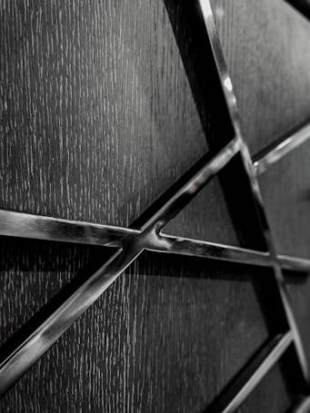 A close-up view of a textured, dark-colored surface intersected by shiny metallic lines forming a grid or network pattern. The metal has a reflective finish, creating a contrast with the matte texture of the background.
