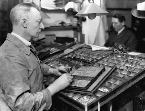 A man is seated at a large wooden desk covered with multiple compartments containing various metal pieces, likely related to typesetting or printing. He is dressed in a work shirt and appears to be focused on a task involving a print block. In the blurred background, another man is sitting at a desk, possibly smoking a pipe.