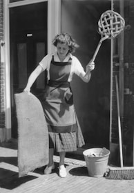 A woman is standing outdoors holding a large wicker carpet beater in one hand and a piece of fabric or mat in the other. She is wearing a striped apron over a plaid skirt and a white blouse. Nearby on the ground, there is a metal bucket and a broom leaning against the wall. The scene appears to be outside a building with brick walls.