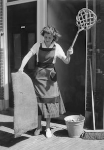 A woman is standing outdoors holding a large wicker carpet beater in one hand and a piece of fabric or mat in the other. She is wearing a striped apron over a plaid skirt and a white blouse. Nearby on the ground, there is a metal bucket and a broom leaning against the wall. The scene appears to be outside a building with brick walls.