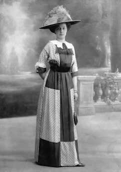 A woman stands in a formal pose wearing a patterned dress from an earlier era, complete with puffed sleeves and a large hat adorned with feathers. The background is a classic studio setting with a painted backdrop that includes what appears to be foliage and a balustrade.