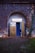 A vintage photo booth is nestled in an ivy-cloaked stone archway. The booth's exterior is metallic with a sign reading 'Foto Automat' perched at the top. A partially open blue curtain reveals a glimpse inside, with part of a stool visible. The surrounding wall is covered in bare, creeping vines, and the ground consists of cobblestones, with patches of green grass visible.