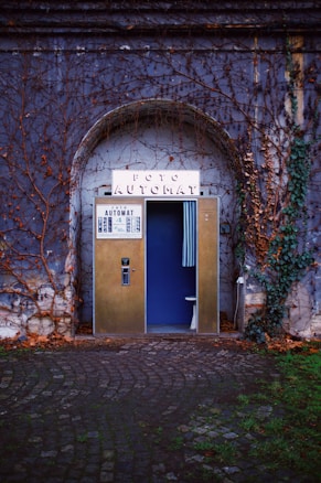 A vintage photo booth is nestled in an ivy-cloaked stone archway. The booth's exterior is metallic with a sign reading 'Foto Automat' perched at the top. A partially open blue curtain reveals a glimpse inside, with part of a stool visible. The surrounding wall is covered in bare, creeping vines, and the ground consists of cobblestones, with patches of green grass visible.
