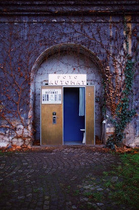 A vintage photo booth is nestled in an ivy-cloaked stone archway. The booth's exterior is metallic with a sign reading 'Foto Automat' perched at the top. A partially open blue curtain reveals a glimpse inside, with part of a stool visible. The surrounding wall is covered in bare, creeping vines, and the ground consists of cobblestones, with patches of green grass visible.