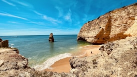 A scenic coastal view featuring towering golden cliffs bordering a secluded beach. The ocean waves gently lap the shore, and a striking rock formation emerges from the water in the distance. The sky is clear with streaks of clouds, and a couple can be seen walking on the sand.