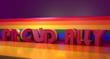 Large 3D letters spelling 'Proud Ally' are presented against a vivid rainbow background. The rainbow pattern extends across the floor and backdrop, casting reflections and enhancing the colorful atmosphere.