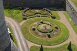 An intricately designed garden featuring symmetrical patterns of greenery and colorful flowers. The garden includes circular arrangements and geometric shapes, bordered by paths and enclosed by stone walls.