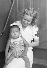 A woman in a nurse's uniform is holding a baby on her lap. The woman has curly hair and is smiling gently at the baby. The baby is wearing a sleeveless outfit and is looking slightly off to the side with a curious expression. They are sitting on a set of wooden steps, creating a simple, intimate scene.